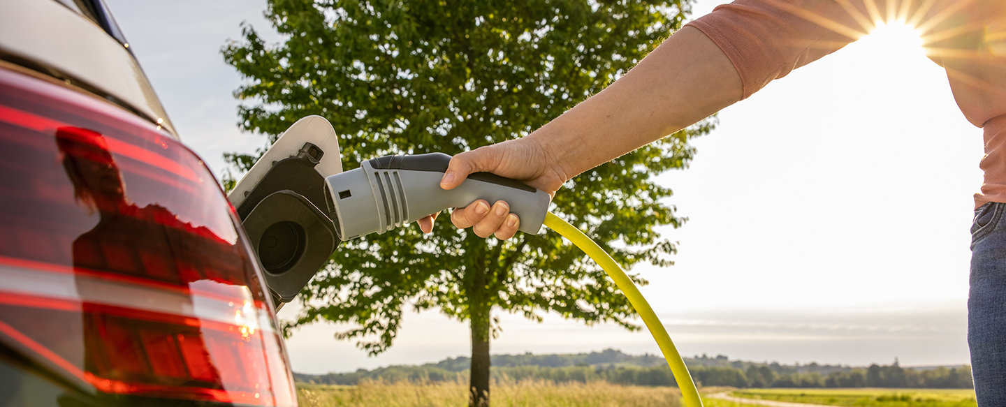 Frau-steckt-ein-Netzkabel-in-ein-Elektroauto-zum-Laden-auf-gruener-Landschaft.jpg